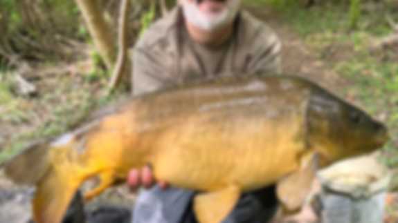 A smiling fisherman poses with a fish he has caught. He has a beard and is wearing a hat.