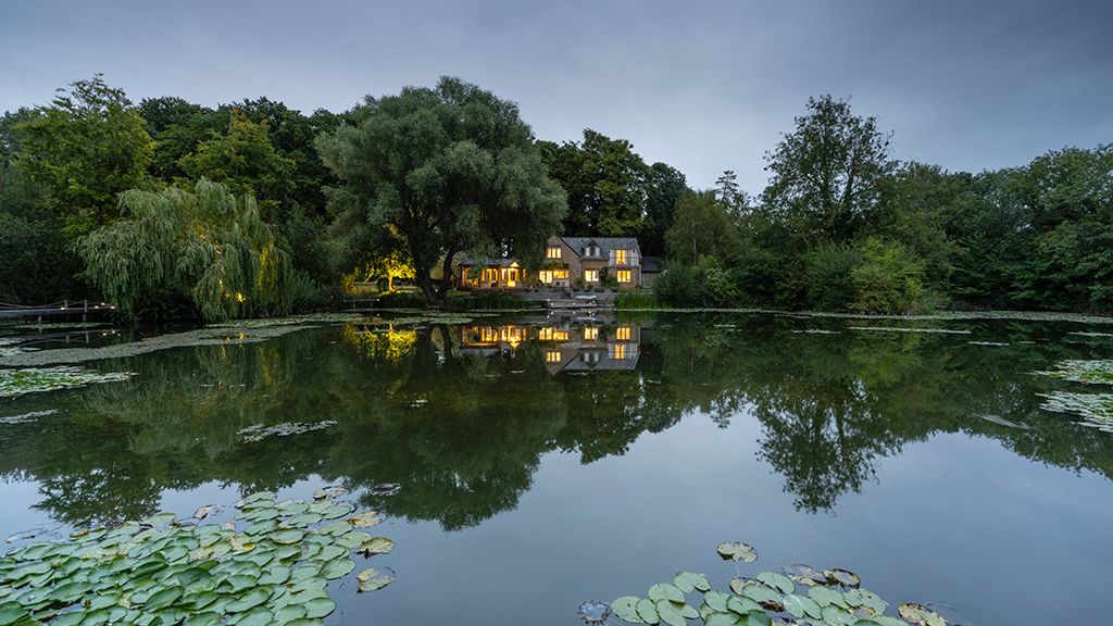 A photo of the house next to the lake from a drone, taken at dusk. The lake and lillies are visible in the foreground.
