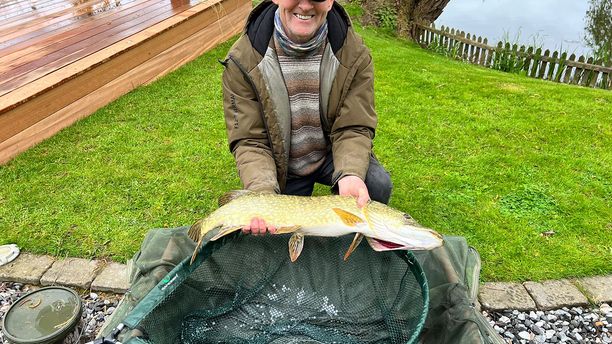 A smiling fisherman poses with a Pike he has caught, whilst kneeling next to the net on the ground.